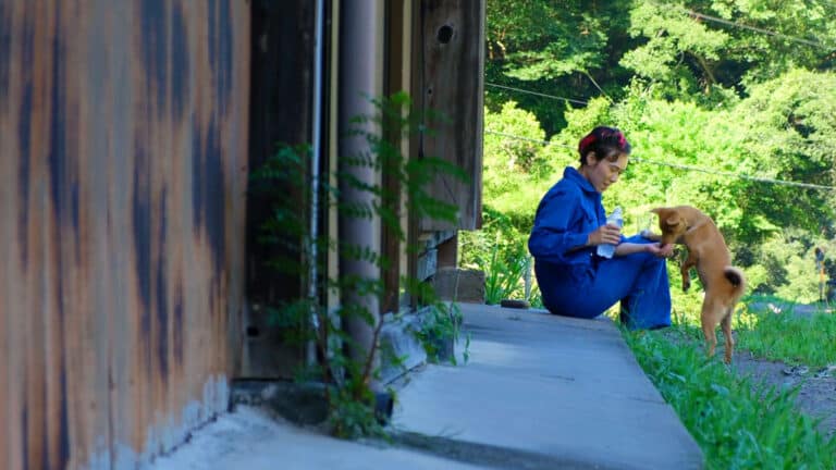 woman sitting next to Japanese house while offering water to dog with curly tail