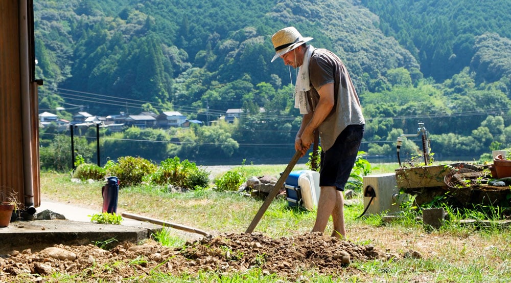 man digging hole with straw hat and sweat-drenched shirt, with blue and green mountain in the background
