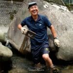 man holding a traditional eel trap in Japan standing in a river looking up happily.